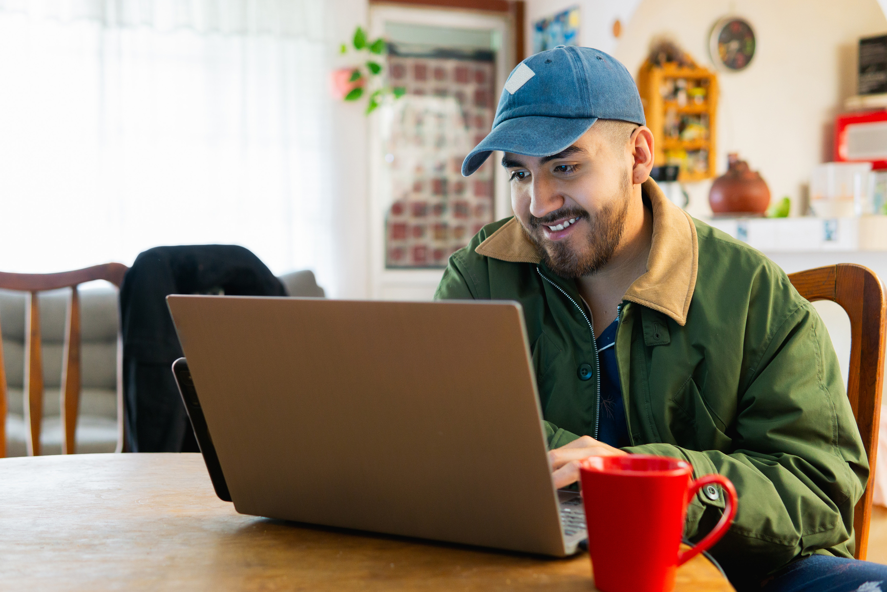 Person wearing a blue cap and green jacket sits at a table and uses a laptop, with a red mug in front of them in a well-lit room.
